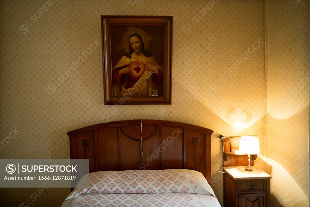 Vintage wall of a bedroom with a picture of the Sacred Heart of Jesus hanging on the headboard of a bed and a bed side table with a lighted lamp. Maó,...