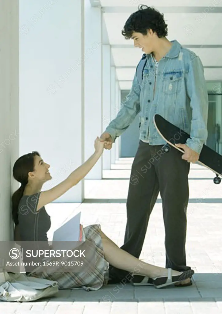 Young man with skateboard reaching hand out to young woman sitting on ground, studying