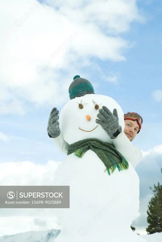 Young man embracing snowman, looking around, portrait