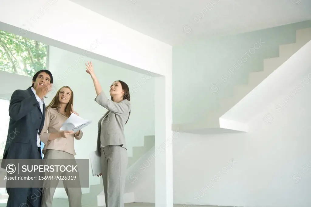 Female real estate agent standing in empty home interior with young couple, full length