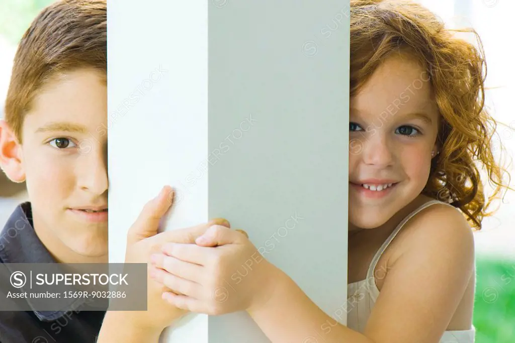 Brother and sister peeking around column at camera, holding hands, smiling