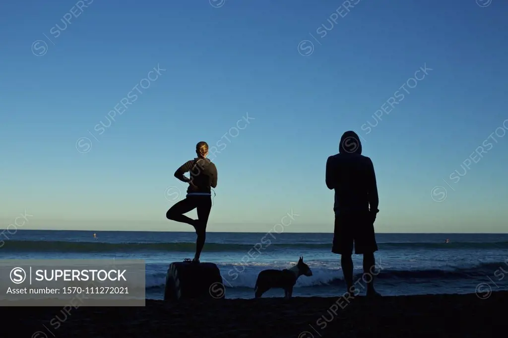 Silhouette young couple with dog practicing yoga on sunrise beach