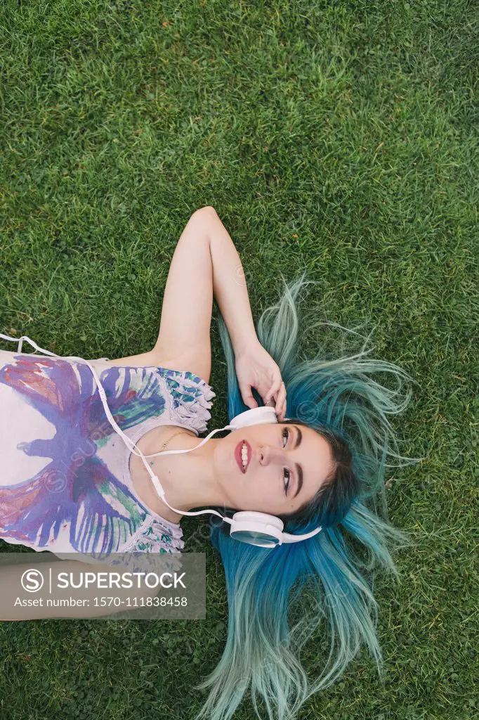 Overhead portrait young woman with blue hair listening to music with headphones, laying in grass