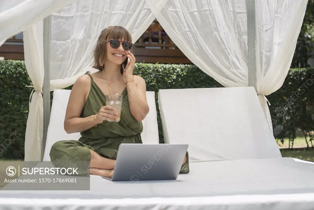 Happy woman talking on smart phone at laptop in cabana