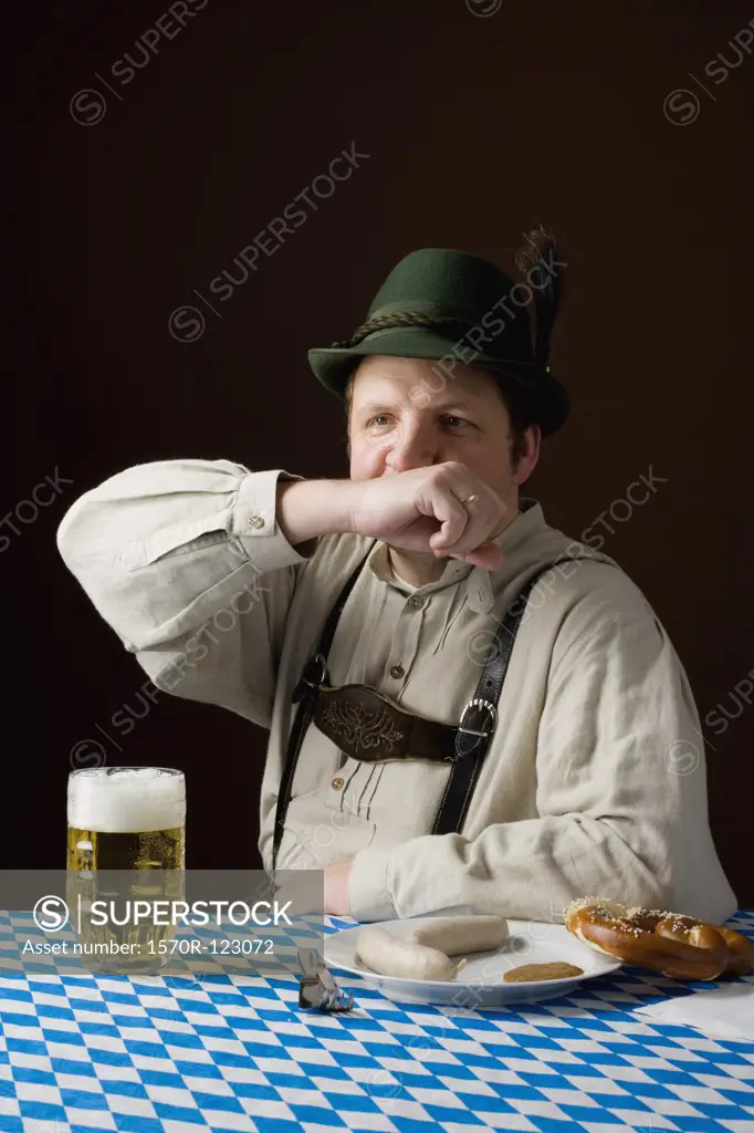 Stereotypical German man in Bavarian costume wiping mouth at a table with a beer and German meal