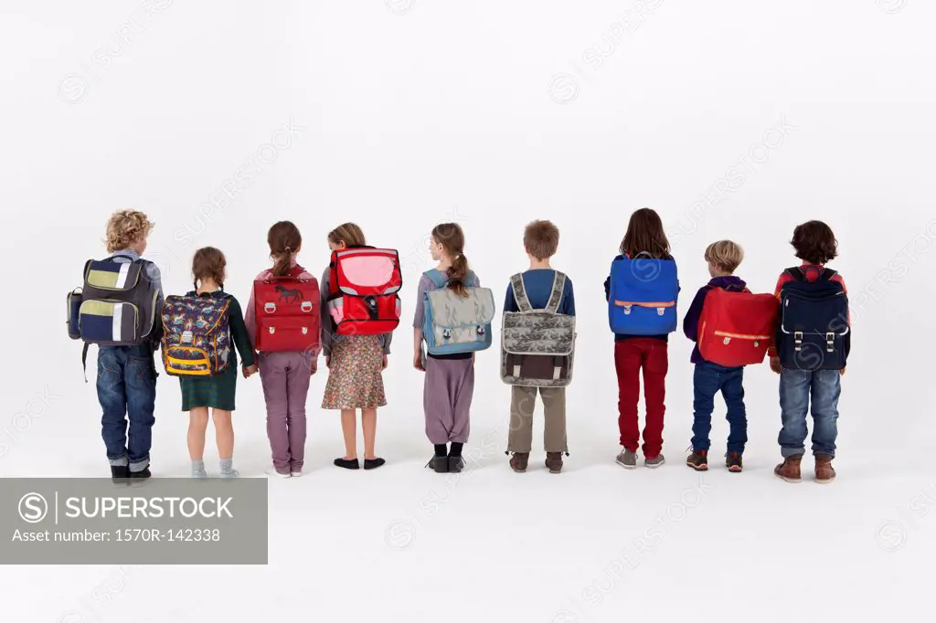 A group of school children wearing backpacks and standing in a row, rear view