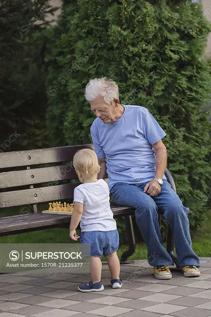 Grandfather playing chess with girl on park bench against tree