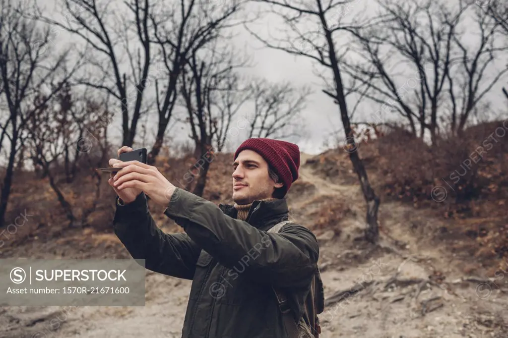 Man with backpack photographing through smart phone while standing against bare trees