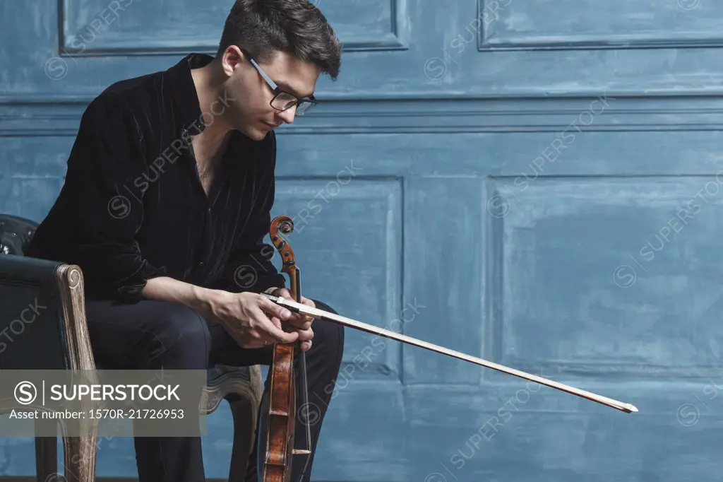 Young man holding violin while sitting on chair by blue wall