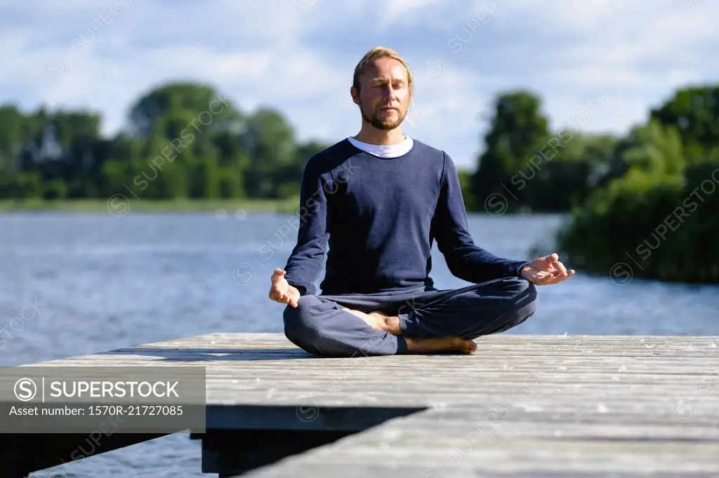 Mature man practicing lotus position on pier by lake against sky