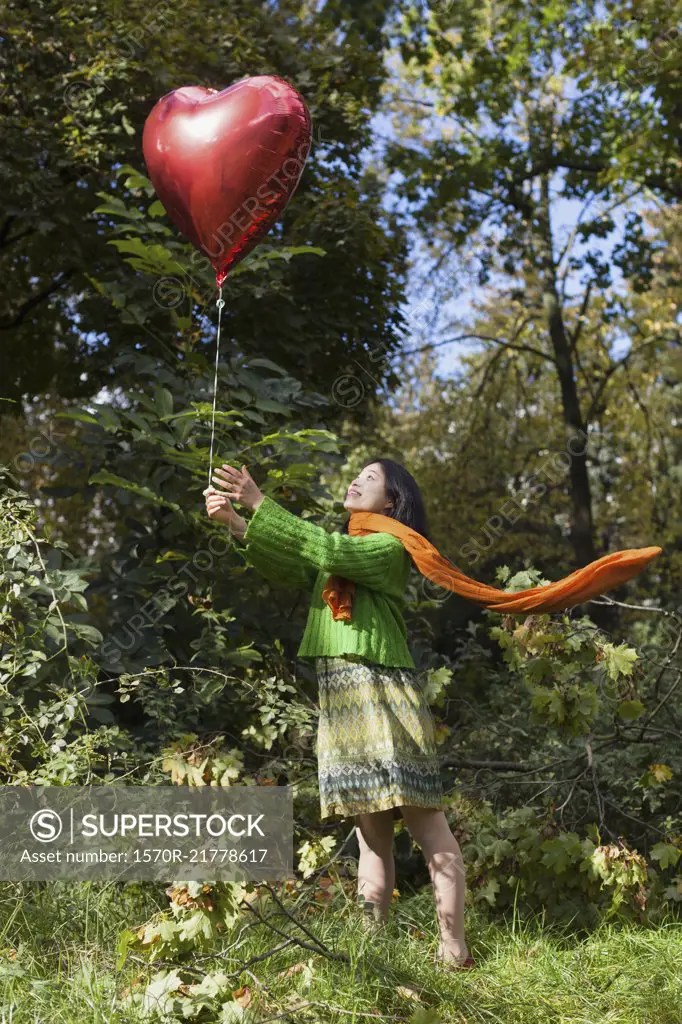 Smiling woman holding red heart shape balloon while standing on field against trees at park