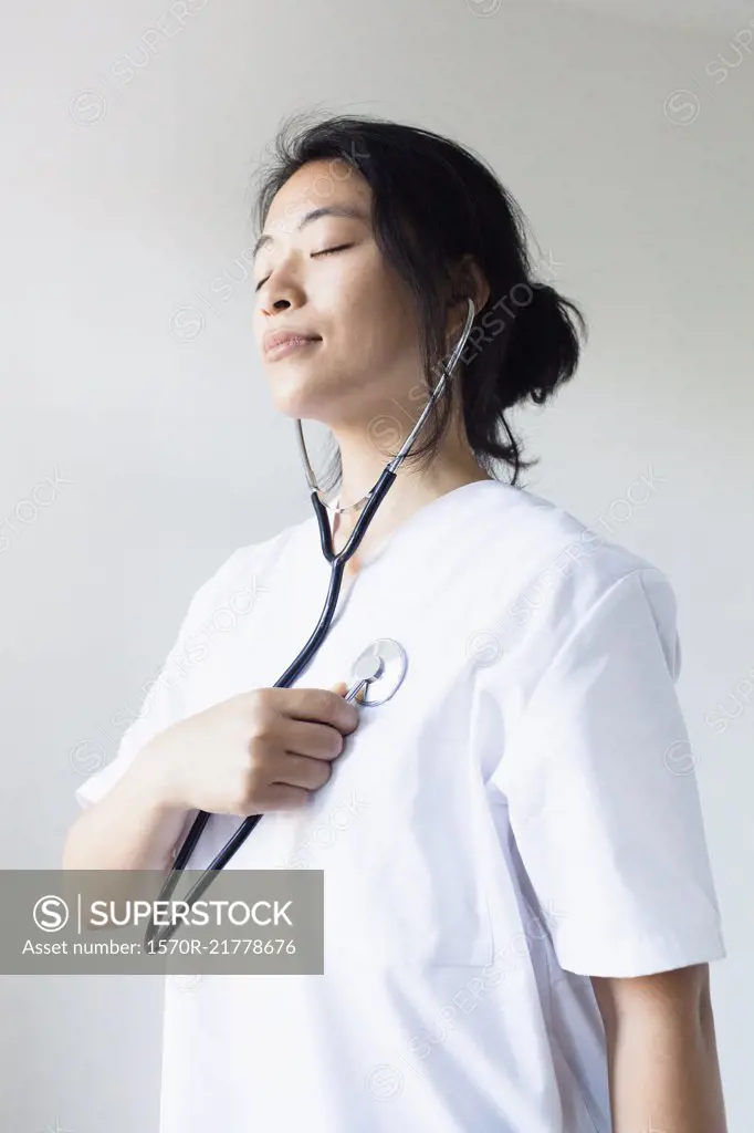 Female doctor listening self heart beat with stethoscope against wall