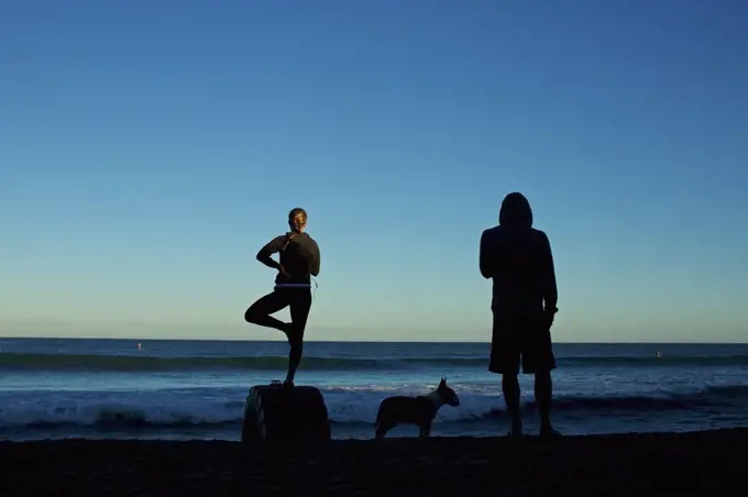 Silhouette young couple with dog practicing yoga on sunrise beach