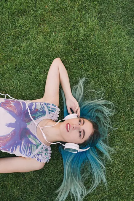 Overhead portrait young woman with blue hair listening to music with headphones, laying in grass