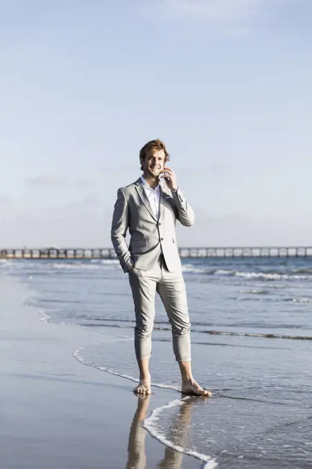 Barefoot businessman talking on smart phone on sunny ocean beach, Los Angeles, California