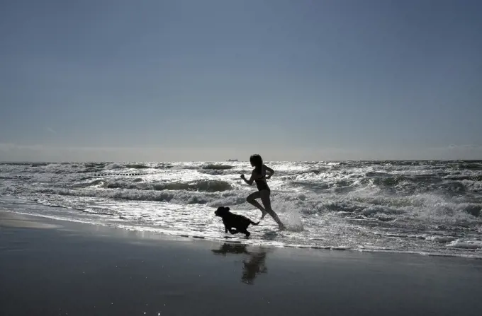 Silhouette girl and pet dog running in sunny, summer ocean surf