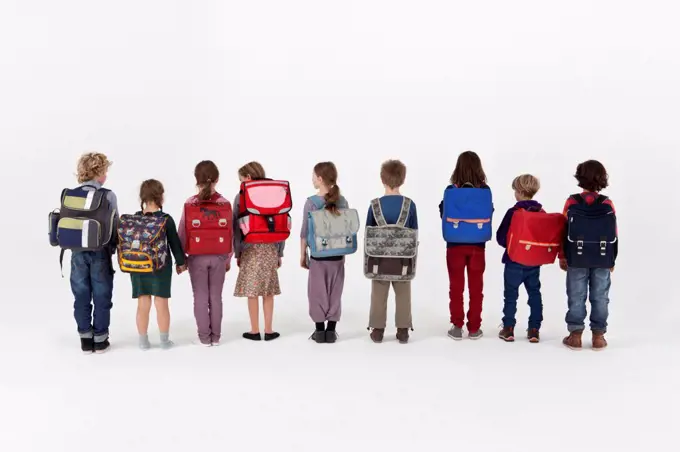 A group of school children wearing backpacks and standing in a row, rear view