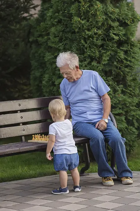 Grandfather playing chess with girl on park bench against tree