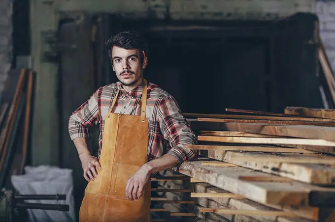 Portrait of carpenter standing by timber stack in workshop