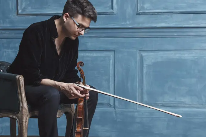 Young man holding violin while sitting on chair by blue wall