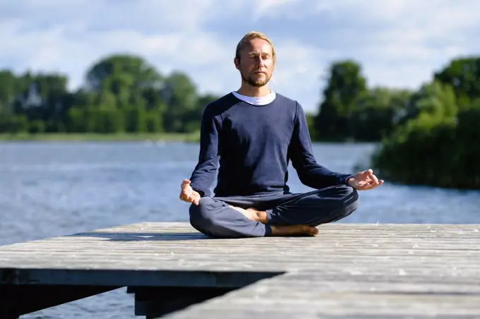 Mature man practicing lotus position on pier by lake against sky