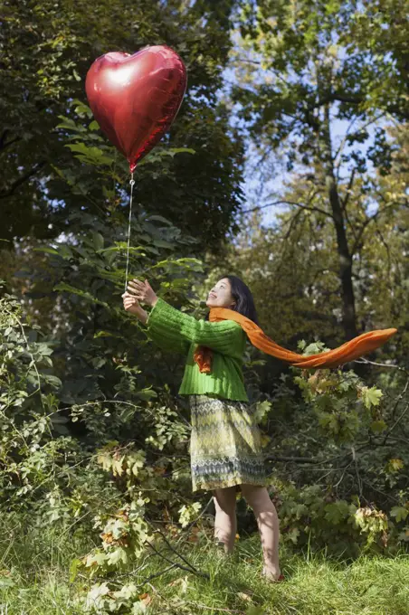 Smiling woman holding red heart shape balloon while standing on field against trees at park