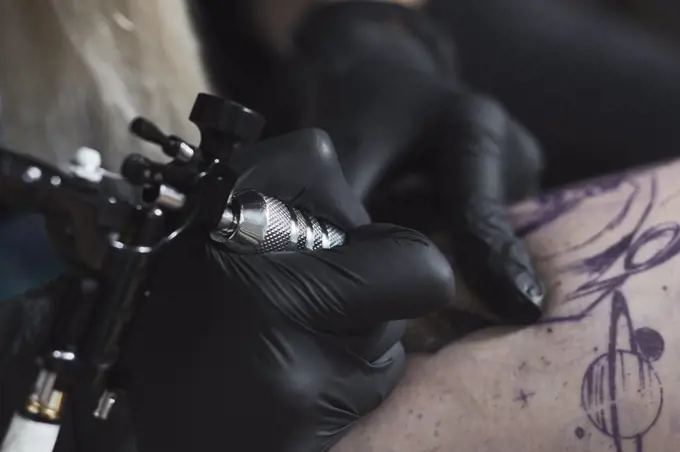 Cropped image of artist wearing gloves tattooing on thigh with machine