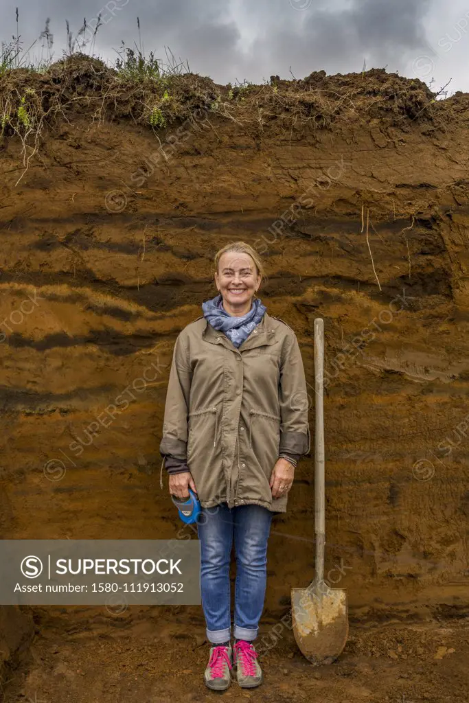 Female with shovel by multiple layers of earth showing deposits of ash from eruptions and volcanic activity, South Coast, Iceland