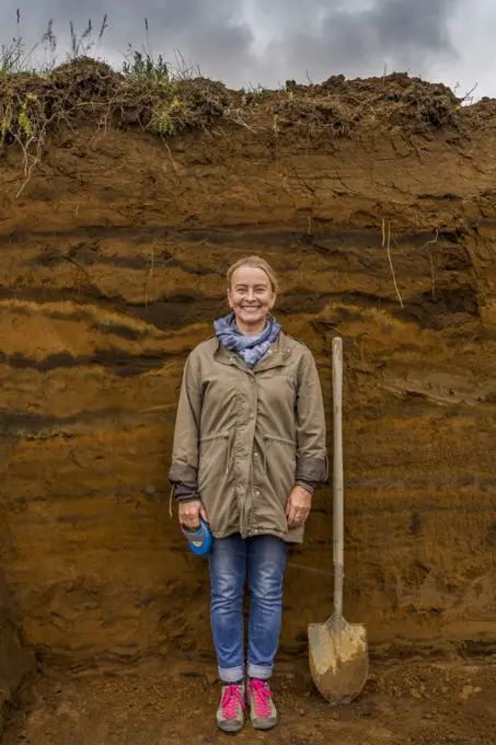 Female with shovel by multiple layers of earth showing deposits of ash from eruptions and volcanic activity, South Coast, Iceland