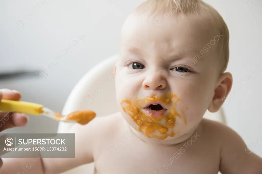 Mother feeding messy baby son with spoon in high chair