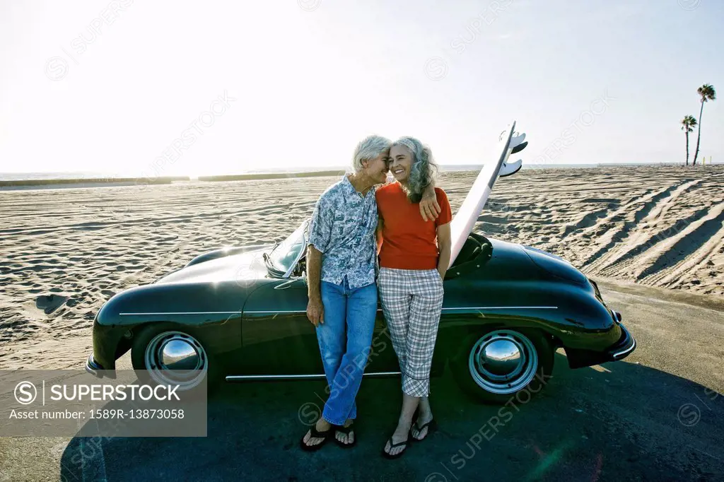 Older Caucasian couple leaning on convertible car with surfboard on beach