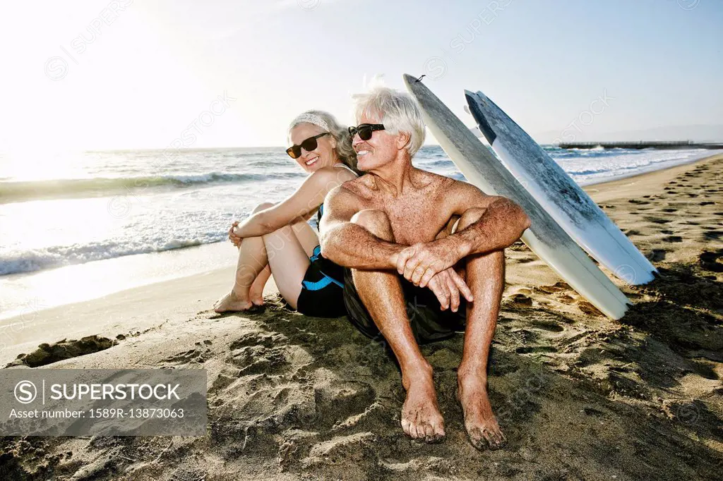 Older Caucasian couple sitting on beach with surfboards