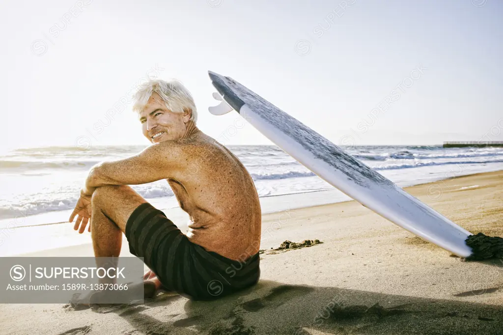 Older Caucasian man sitting on beach with surfboard