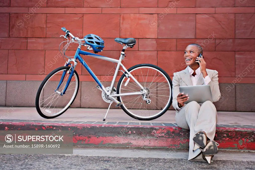 Businesswoman sitting on curb using digital tablet and cell phone