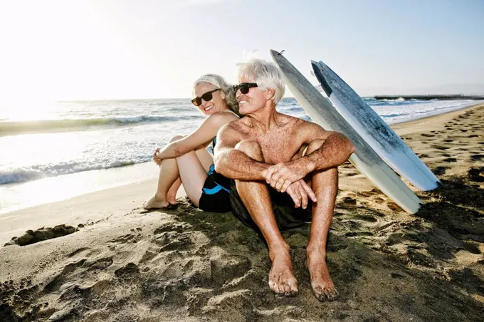 Older Caucasian couple sitting on beach with surfboards