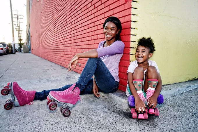 Black mother and daughter wearing roller skates sitting on sidewalk