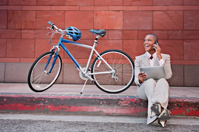 Businesswoman sitting on curb using digital tablet and cell phone