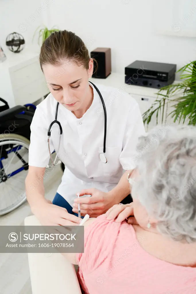 Cheerful young woman doctor giving vaccine injection to elderly woman at home.