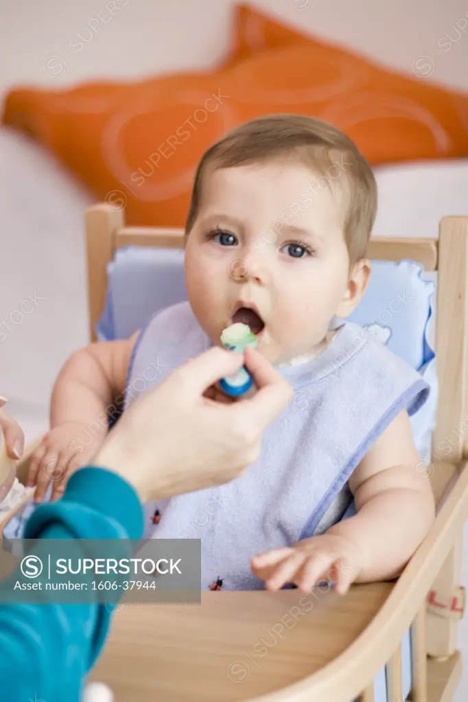 Close-up on womans's hand feeding little boy, seated in high chair, small plastic pot