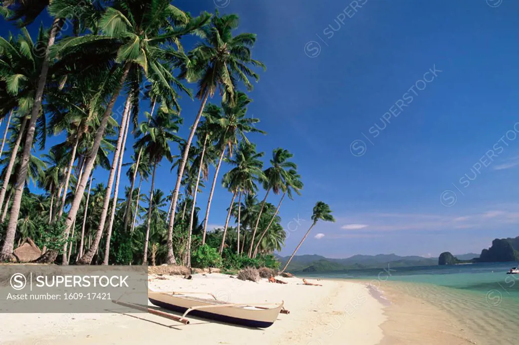 Philippines, Palawan, Bascuit Bay, El Nido, Couple Sunbathing on Beach with Outrigger Boat in Foreground