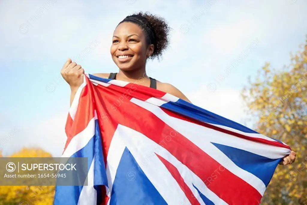 London, UK. Smiling young woman holding British flag against trees and sky