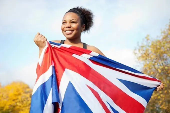 London, UK. Smiling young woman holding British flag against trees and sky