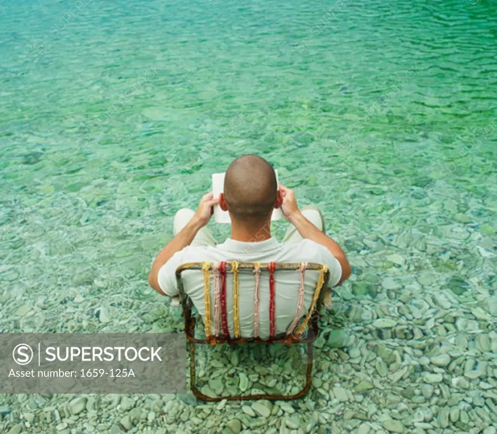 High angle view of a young man sitting in chair in water and reading a book