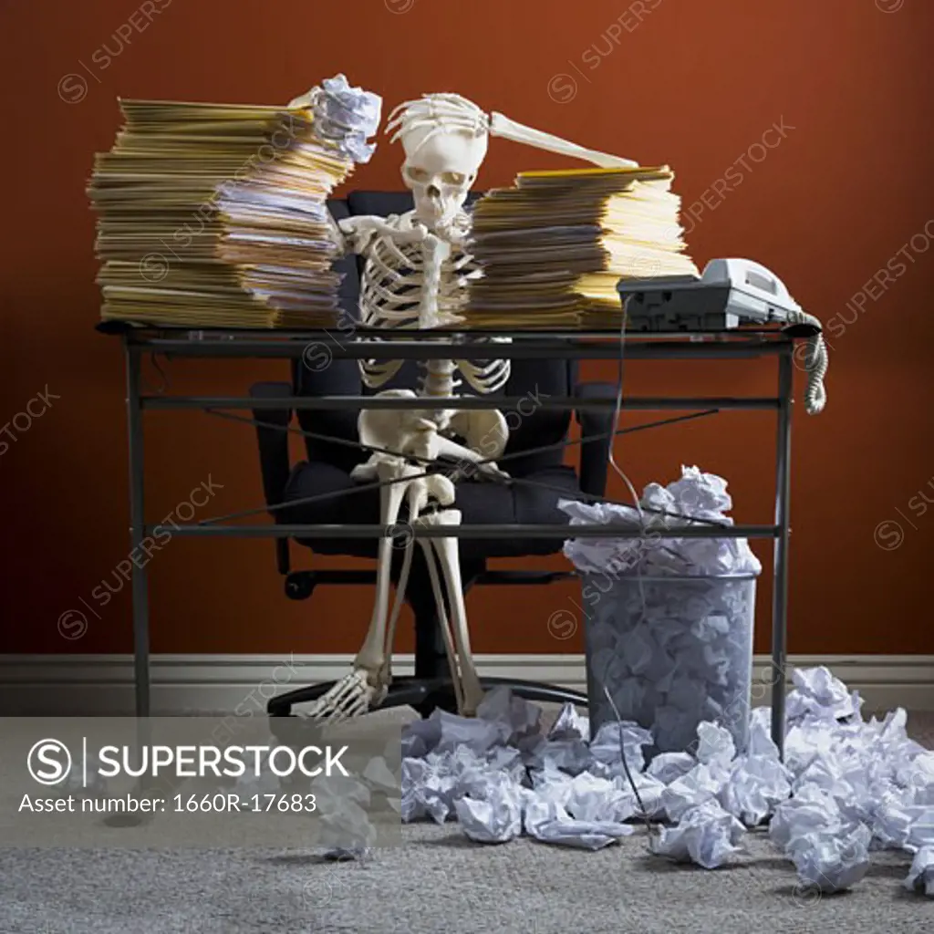 Skeleton sitting at desk with stacks of paperwork and crumpled papers