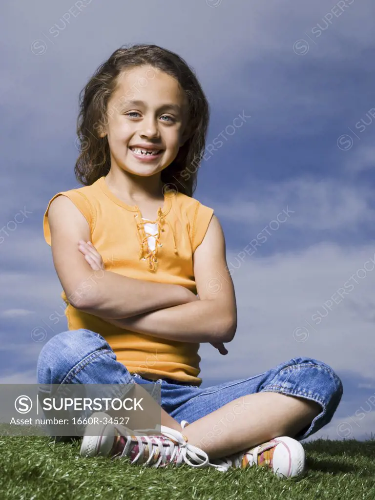 Girl sitting cross legged on grass outdoors with arms crossed smiling