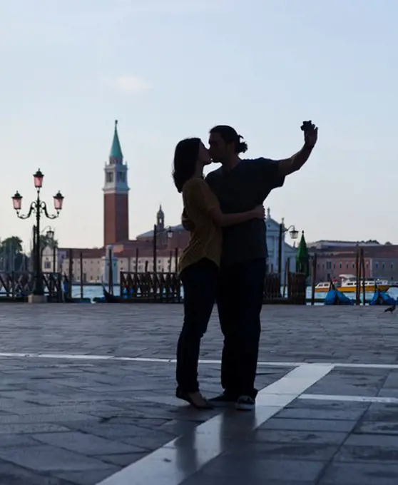 Italy, Venice, Young couple kissing and photographing self, San Giorgio Maggiore church in background