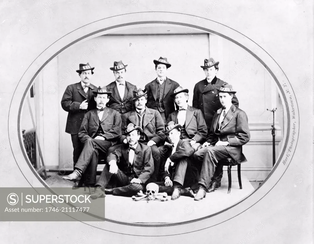 Photograph shows ten men posed, seated and standing, wearing hats with 'KKK' in large letters, and with a skull and bones arranged on the floor in front of them. Ku Klux Klan, Watertown Division, photographer, Watertown, New York.