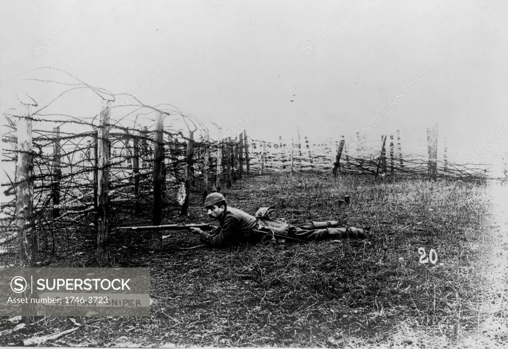 World War I 1914-1918: German sniper wearing a pickelhelm, lying on the ground behind barbed wire entanglements, rifle at the ready.