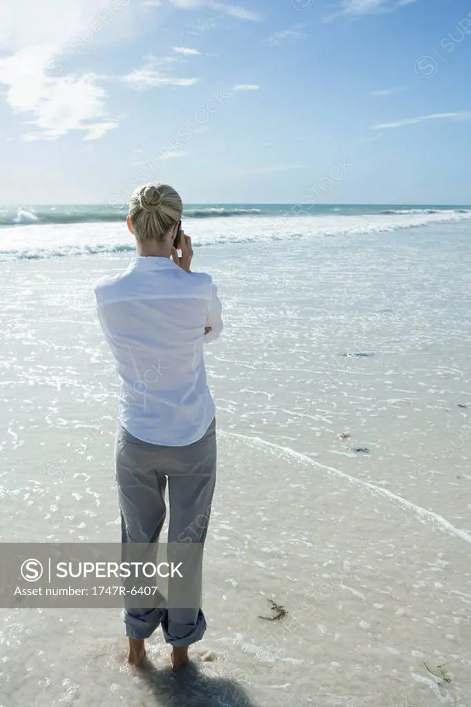 Woman standing in surf, using cell phone, rear view
