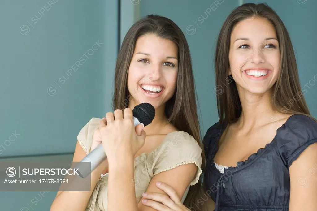 Teenage twin sisters side by side, both smiling, one holding microphone